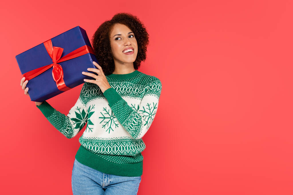 sonriente y curiosa mujer afroamericana en suéter de invierno sacudiendo caja de regalo azul aislado en rojo - Foto, Imagen