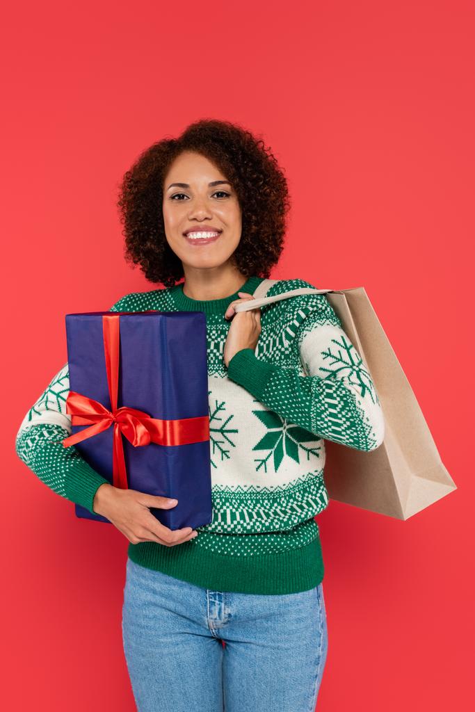 mulher americana africana feliz em camisola quente segurando saco de compras e caixa de presente azul isolado no vermelho - Foto, Imagem
