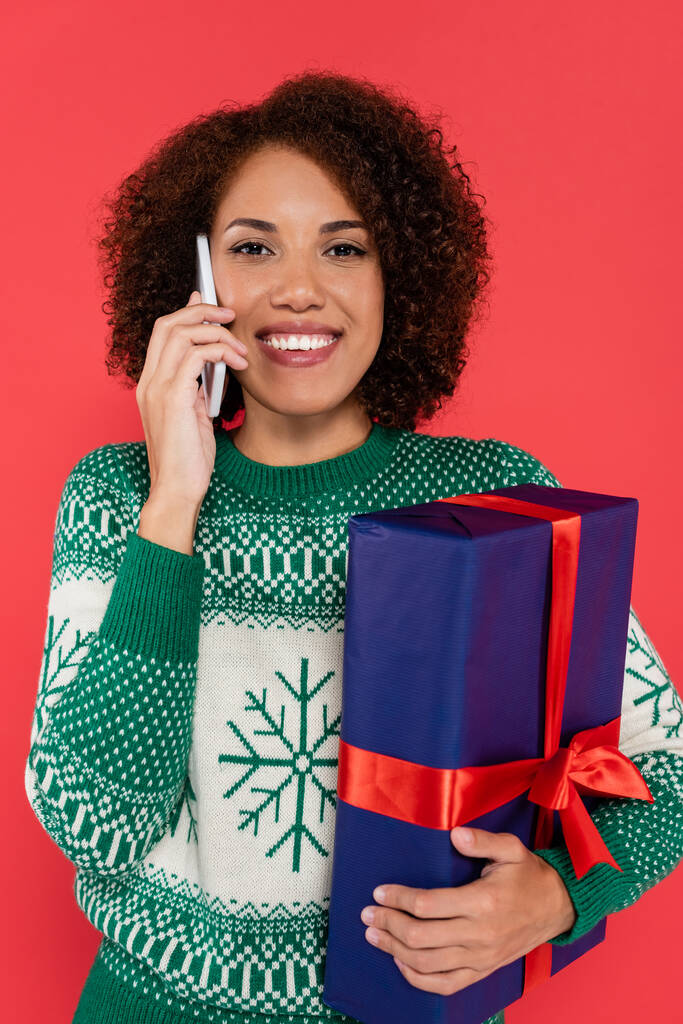 cheerful african american woman with blue gift box talking on mobile phone isolated on red - Photo, Image