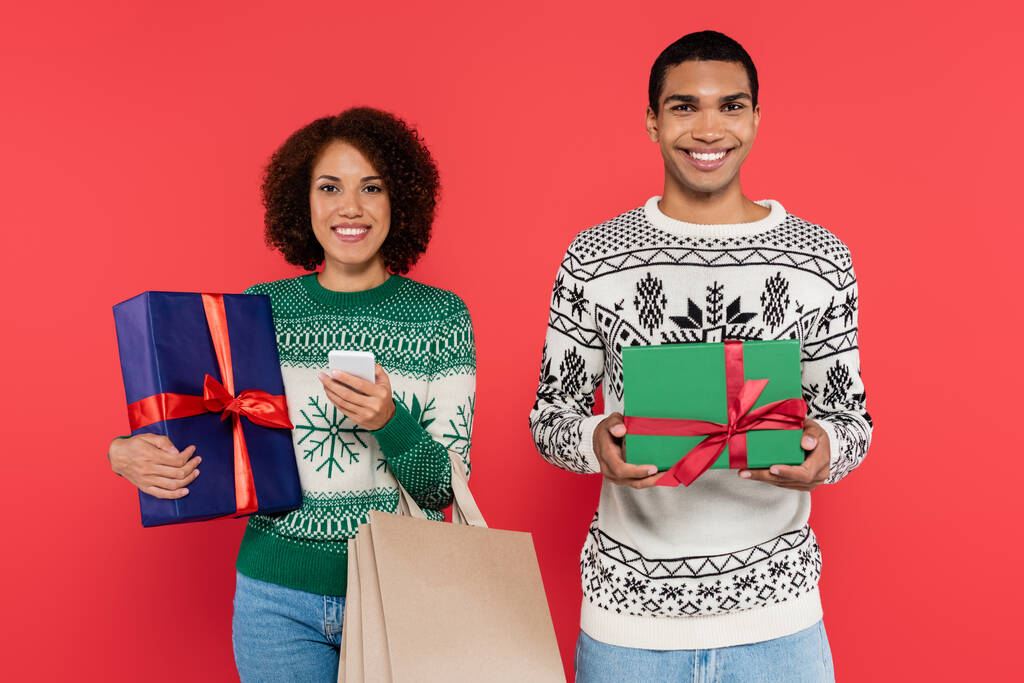 afro-amerikai nő gazdaság okostelefon bevásárló táskák és ajándékdoboz közelében boldog férfi jelen elszigetelt piros - Fotó, kép