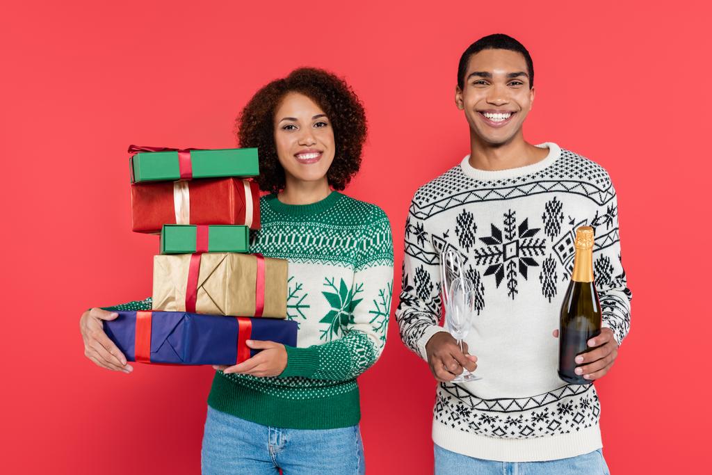 усміхнена афроамериканська жінка з різнокольоровими подарунковими коробками біля чоловіка з шампанським ізольовано на червоному
 - Фото, зображення