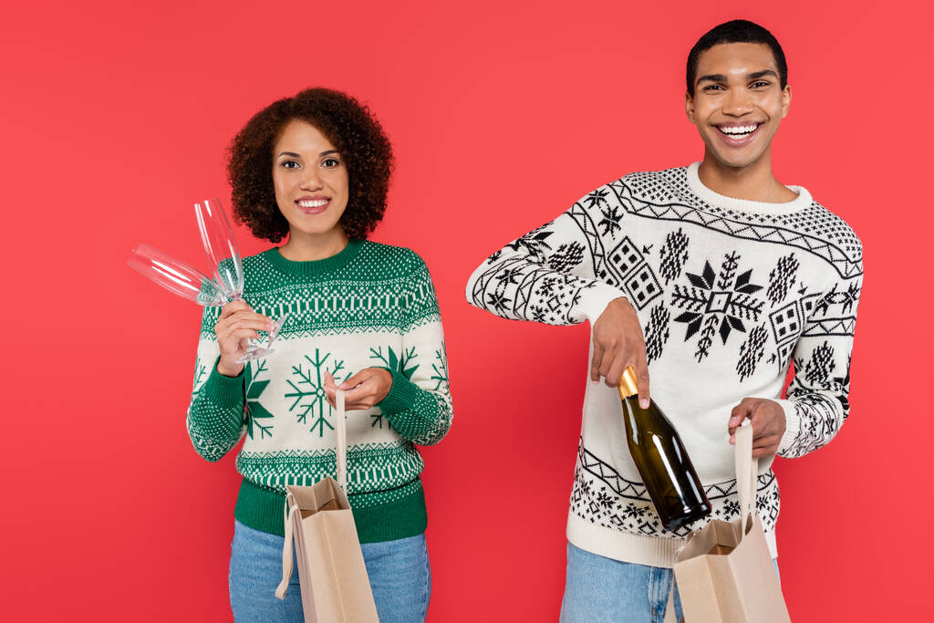 Alışveriş çantaları ve şampanya taşıyan mutlu Afro-Amerikan çifti kırmızı ışıkta izole edilmiş kameraya bakıyor. - Fotoğraf, Görsel