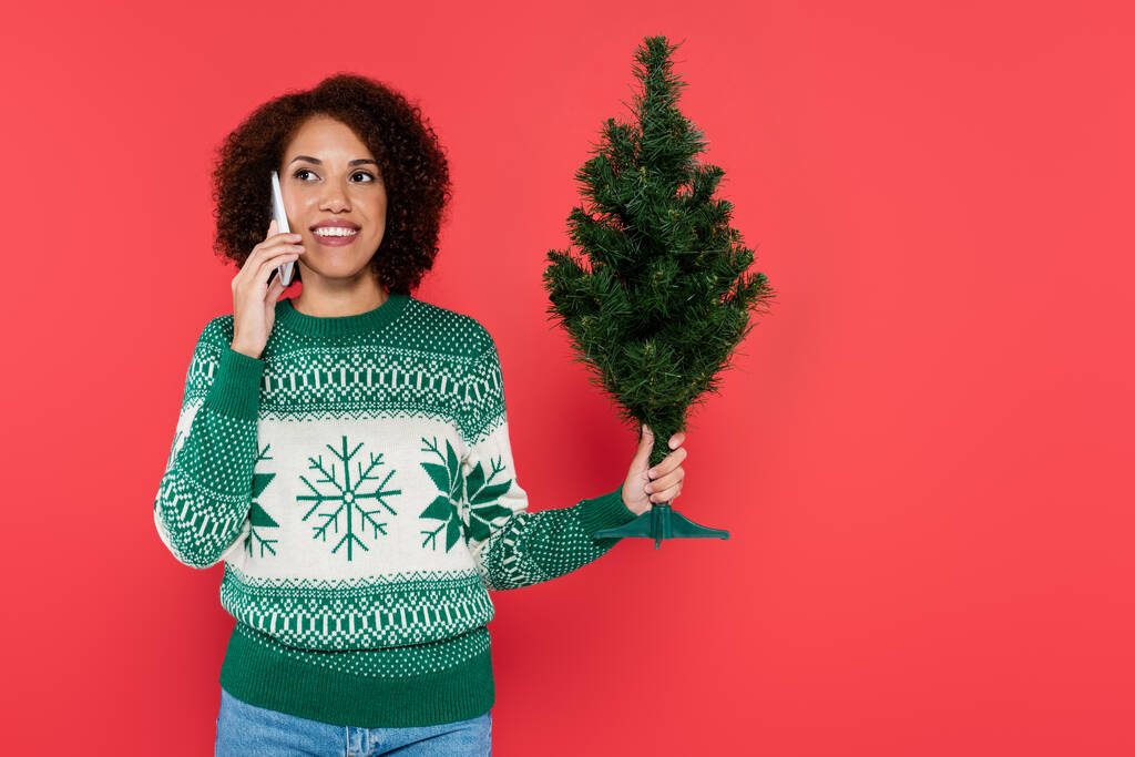 vidám afro-amerikai nő fehér pulóverben gazdaság kis karácsonyfa és beszél okostelefon elszigetelt piros - Fotó, kép