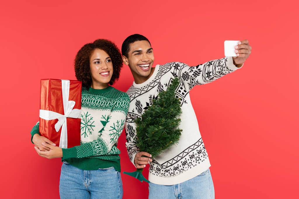 veselý africký Američan muž s malým vánočním stromečkem přičemž selfie s přítelkyní drží dárek izolované na červené - Fotografie, Obrázek