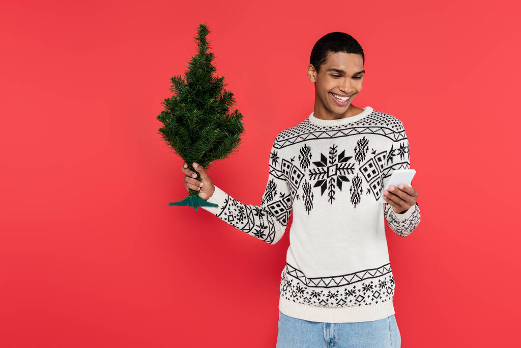 homme afro-américain souriant en pull avec motif d'hiver tenant petit arbre de Noël et téléphone portable isolé sur rouge - Photo, image
