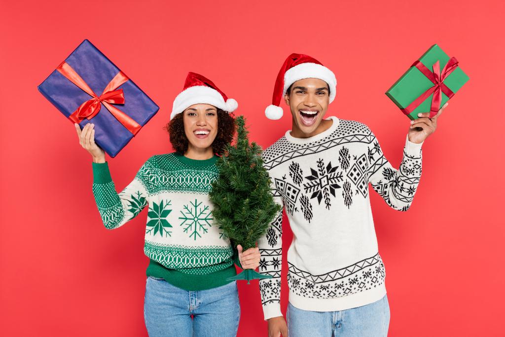 excited african american couple in santa hats and warm sweaters holding green and blue gift boxes isolated on red - Photo, Image