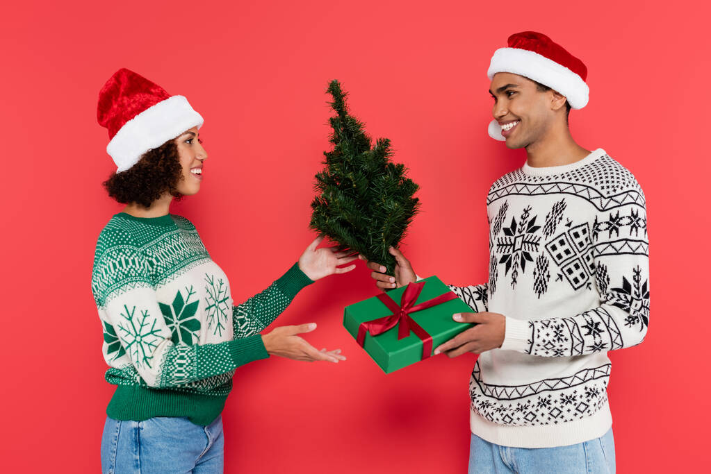 lächelnder afrikanisch-amerikanischer Mann mit Weihnachtsmütze überreicht Geschenk und kleinen Weihnachtsbaum an zufriedene Freundin isoliert auf rot - Foto, Bild