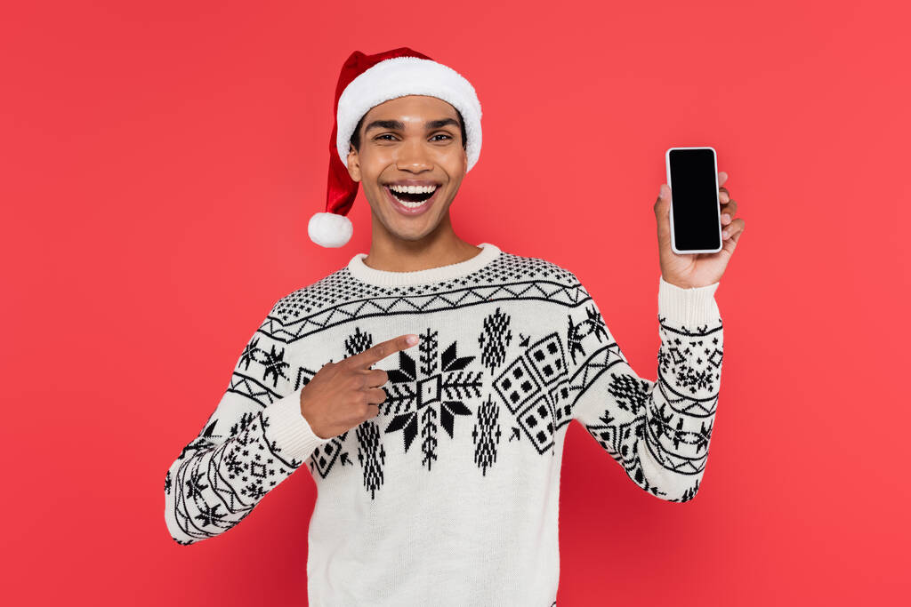 hombre afroamericano riendo en sombrero de santa señalando el teléfono móvil con la pantalla en blanco aislado en rojo - Foto, Imagen