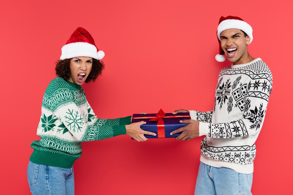excité couple afro-américain dans santa chapeaux crier tout en tenant boîte cadeau bleu isolé sur rouge - Photo, image
