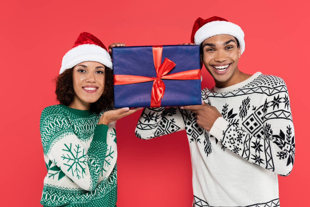 Ευτυχισμένο ζευγάρι Αφροαμερικανών με καπέλα και ζεστά πουλόβερ που κρατάνε ένα μεγάλο κουτί δώρου απομονωμένο στο κόκκινο. - Φωτογραφία, εικόνα