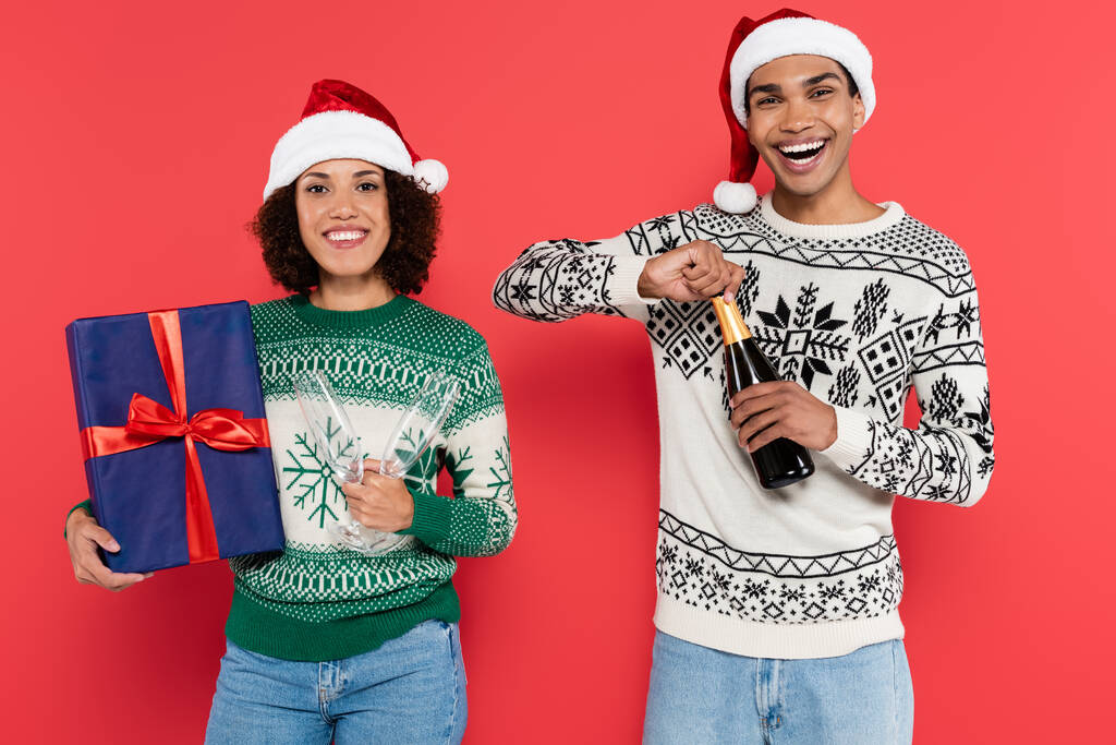 joyeux homme afro-américain ouverture champagne près de petite amie avec boîte cadeau bleu isolé sur rouge - Photo, image