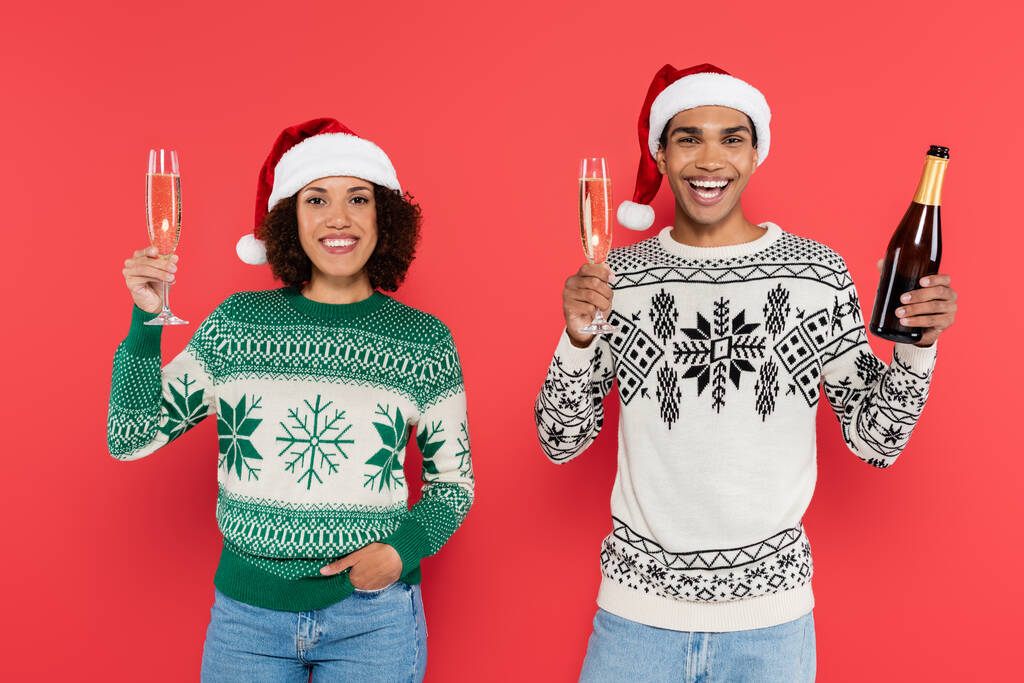gioiosa coppia afroamericana in cappelli di Babbo Natale in possesso di bicchieri di champagne e guardando la fotocamera isolata sul rosso - Foto, immagini