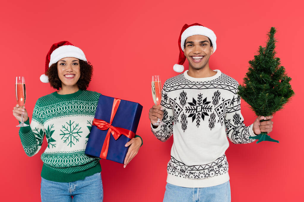 fröhliches afrikanisch-amerikanisches Paar mit Champagnergläsern mit Weihnachtsbaum und blauer Geschenkschachtel isoliert auf rot - Foto, Bild