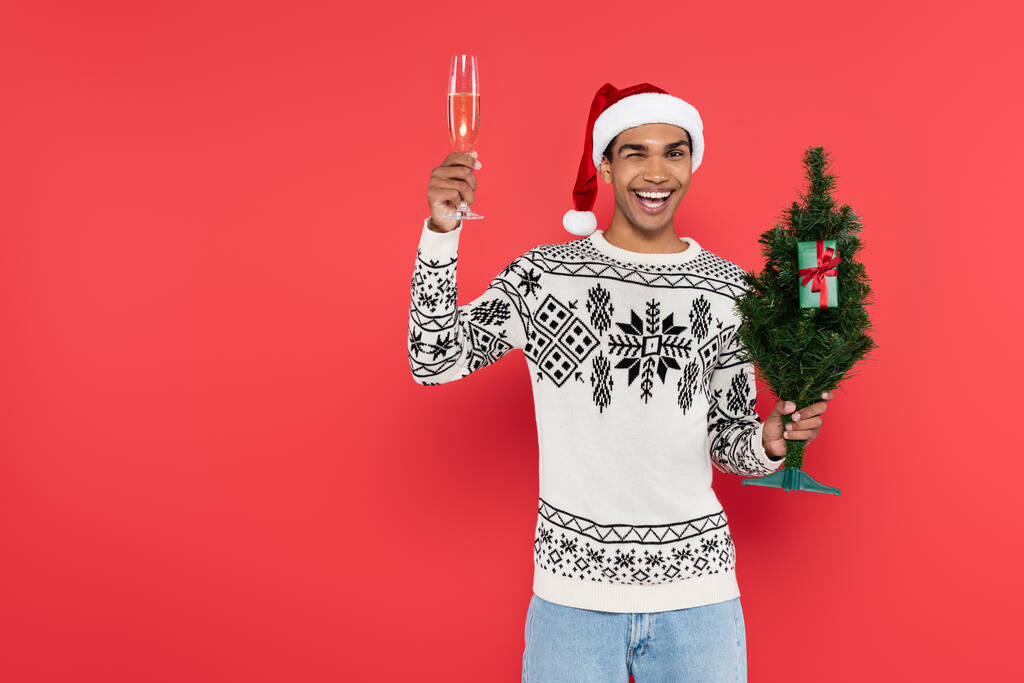 vrolijk Afrikaans amerikaanse man met kerstboom en champagne glas knipperen op camera geïsoleerd op rood - Foto, afbeelding