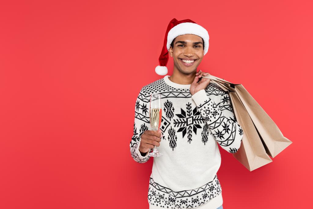 african american man in santa hat holding shopping bags and champagne glass while smiling at camera isolated on red - Photo, Image