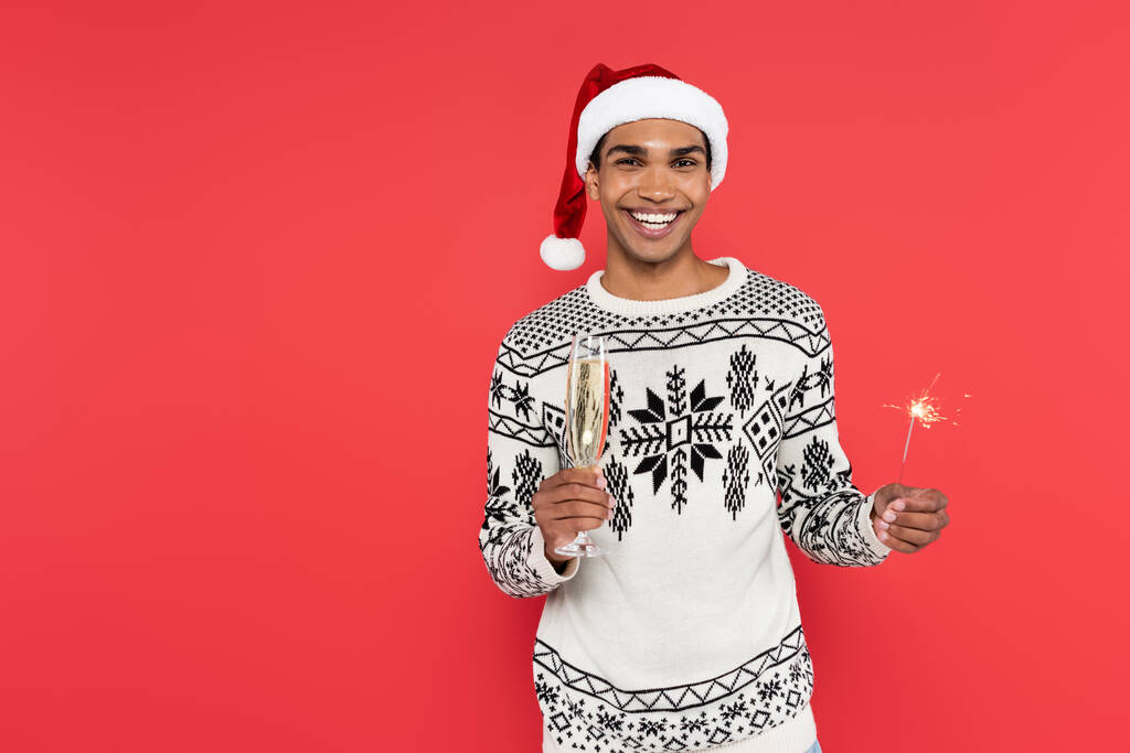 cheerful african american man in santa hat and sweater with winter pattern holding champagne glass and sparkler isolated on red - Photo, Image