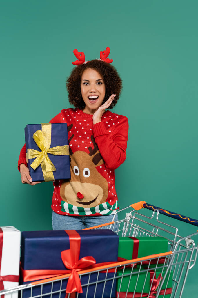 amazed african american woman in red sweater with reindeer holding present near shopping cart isolated on green - Photo, Image