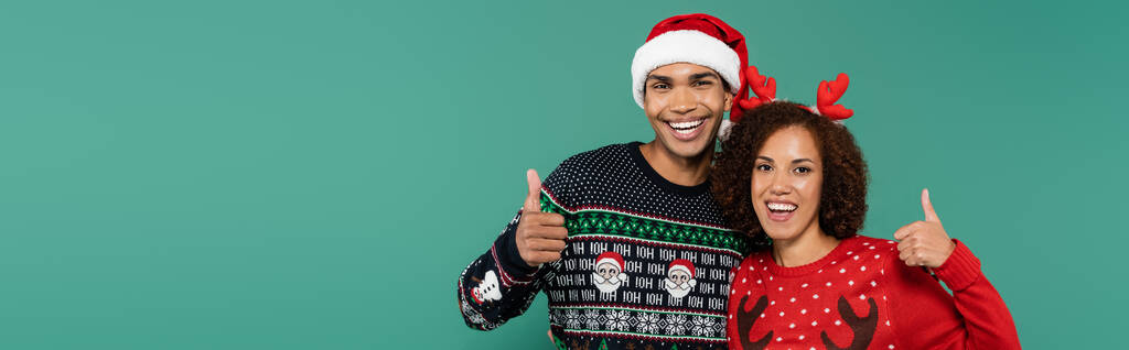 smiling african american woman in deer horns headband and man in santa hat showing thumbs up isolated on green, banner - Photo, Image