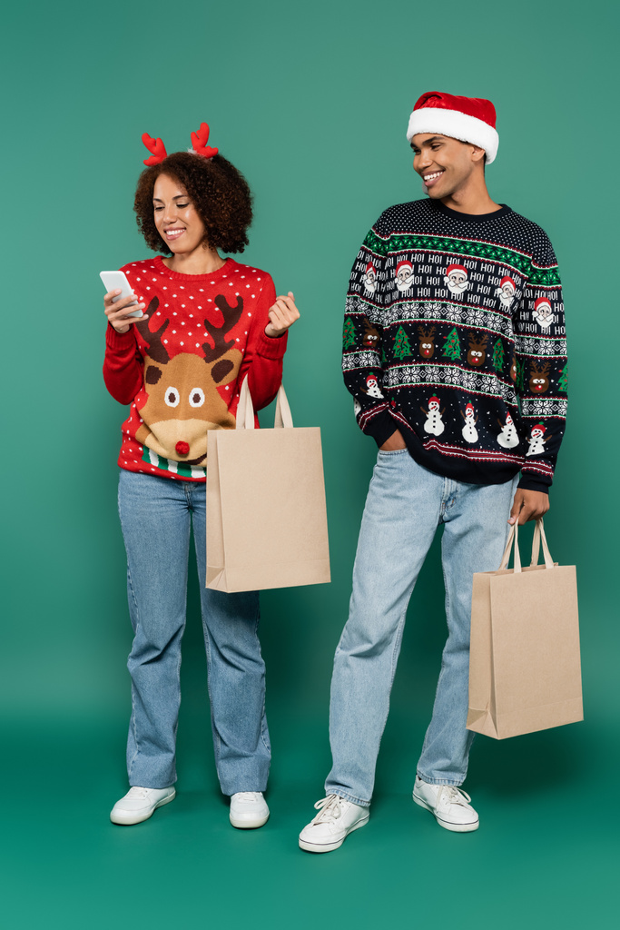 クリスマスの衣装を着たアフリカ系アメリカ人女性はサンタ・ハットの笑顔の男の近くでスマートフォンを見て - 写真・画像