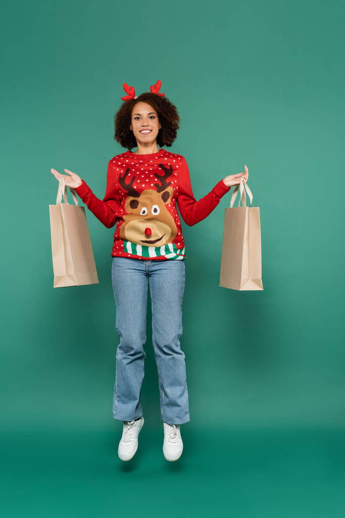 glücklich afrikanisch-amerikanische Frau im Pullover mit Hirsch und roten Hörnern Stirnband schwebt mit Einkaufstaschen auf grünem Hintergrund - Foto, Bild