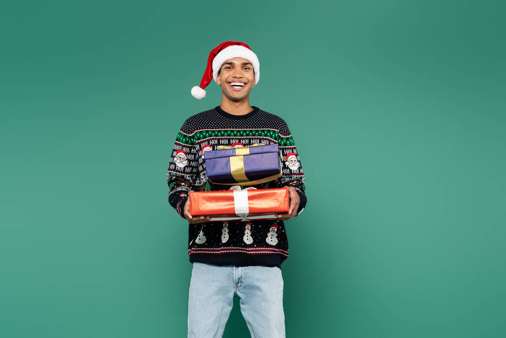 animado afro-americano homem em santa chapéu e inverno suéter segurando Natal apresenta isolado no verde - Foto, Imagem