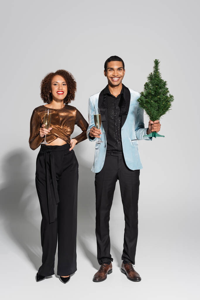 повна довжина елегантної і стильної афроамериканської пари з шампанським і маленькою різдвяною ялинкою на сірому фоні
 - Фото, зображення