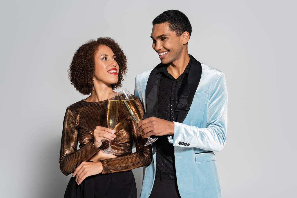 jovem casal afro-americano em roupas elegantes clinking taças de champanhe e sorrindo um para o outro isolado em cinza - Foto, Imagem