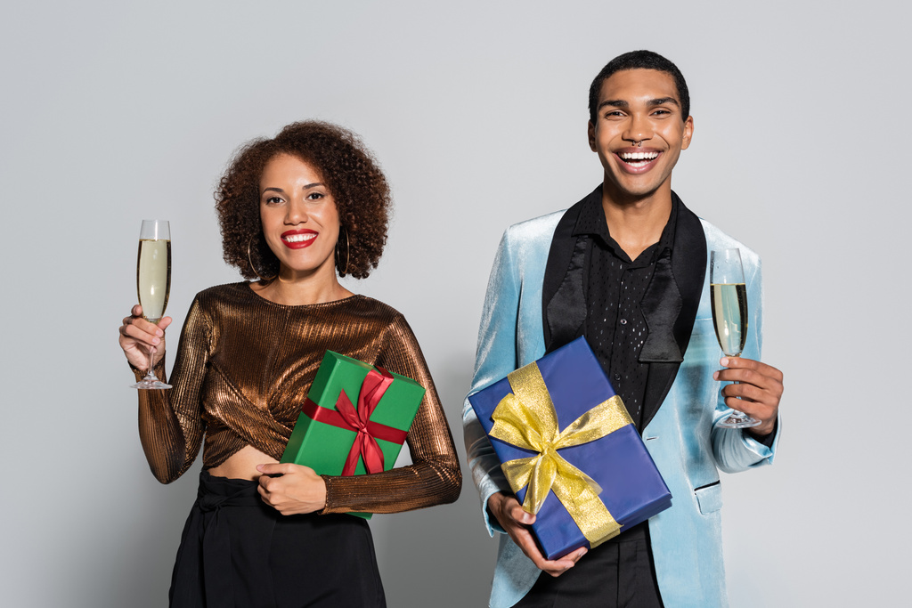 boldog afro-amerikai pár elegáns ruhákban kezében pezsgő és ajándékdobozok elszigetelt szürke - Fotó, kép