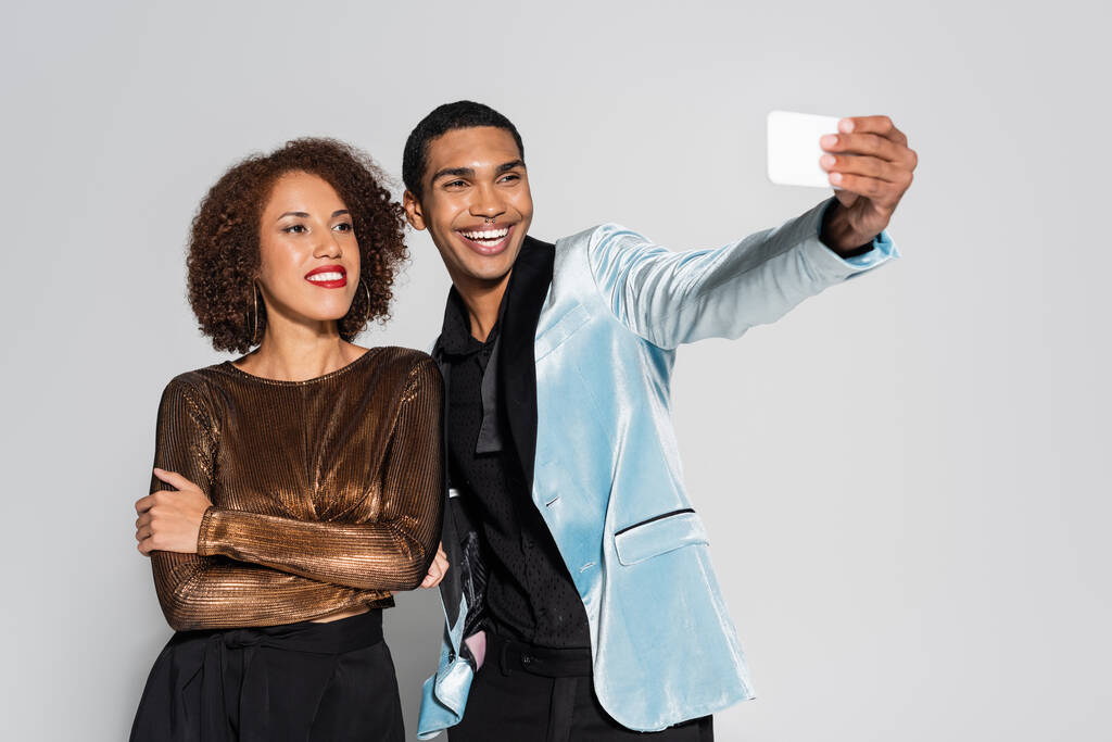 Χαρούμενος Αφροαμερικάνος που βγάζει selfie με την κομψή φιλενάδα του να στέκεται με σταυρωμένα χέρια απομονωμένη στο γκρι - Φωτογραφία, εικόνα