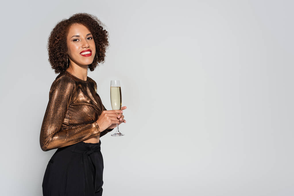 curly african american woman in elegant shiny blouse smiling at camera while standing with champagne isolated on grey - Photo, Image