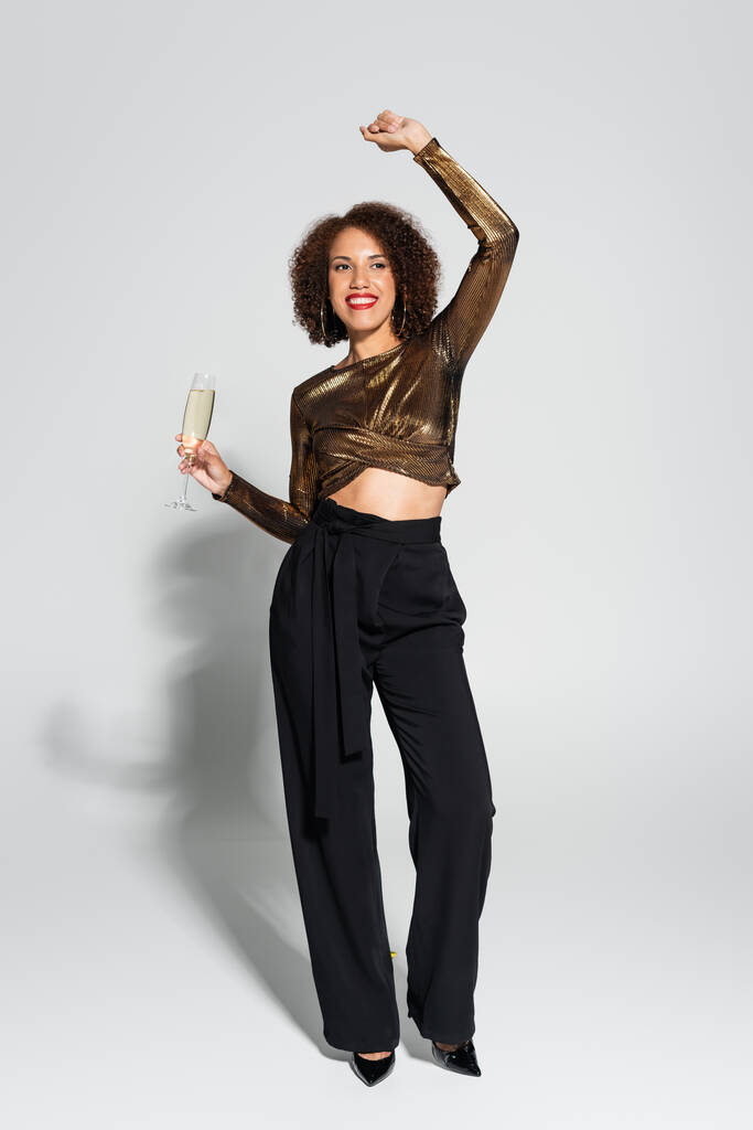 full length of african american woman in brown blouse and black pants dancing with champagne glass on grey background - Photo, Image