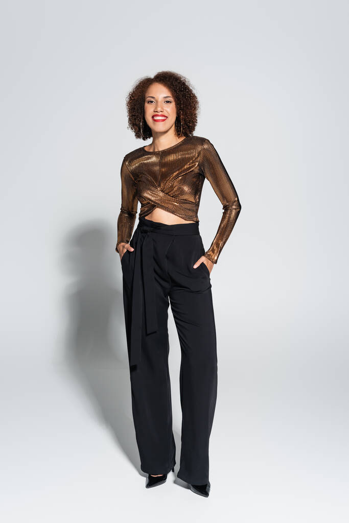 Кудрявая африканская американка в коричневой и блестящей блузке позирует с руками в кармане черных брюк на сером фоне - Фото, изображение