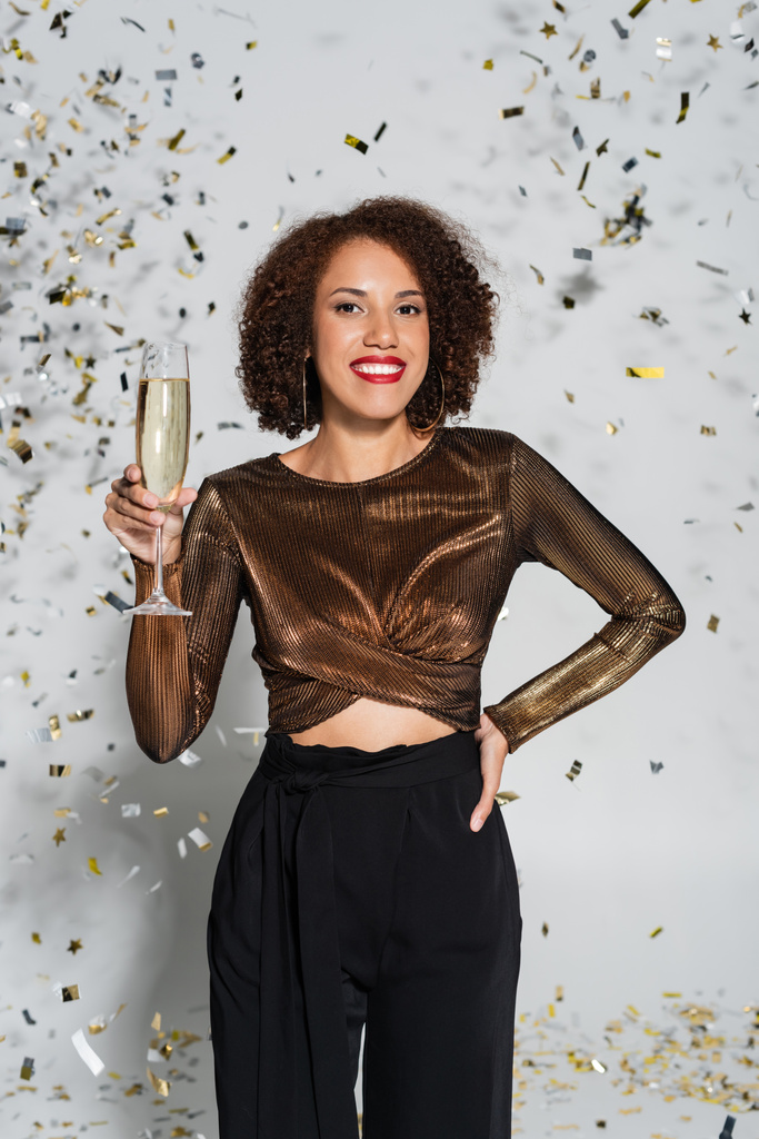 Радісна афро-американська жінка стоїть з рукою на бедрах і шампанському склі біля конфетті на сірому фоні. - Фото, зображення
