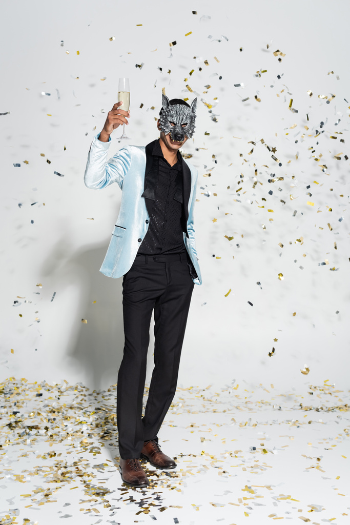 comprimento total de homem americano africano elegante em tostagem máscara lobo com champanhe perto confetti no fundo cinza - Foto, Imagem