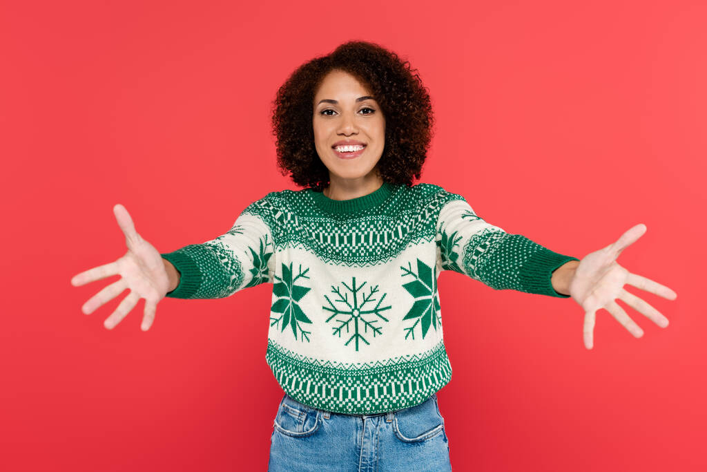 glückliche afrikanisch-amerikanische Frau im Winterpullover mit grünem Muster, die mit ausgestreckten Händen auf rotem Grund in die Kamera blickt - Foto, Bild
