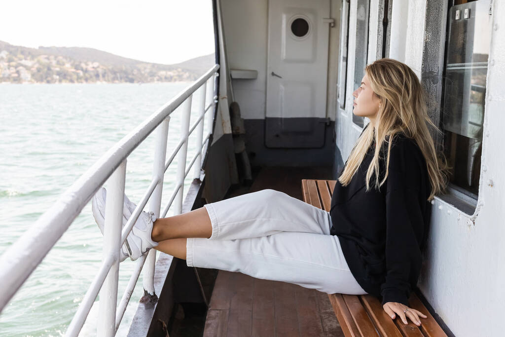 blond vrouw in zwart trui zitten op bank terwijl kijken naar de zee van veerboot kruising bosporus in Istanbul  - Foto, afbeelding