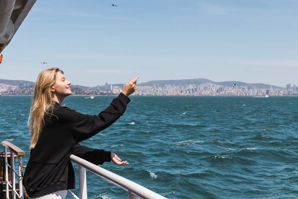 szczęśliwa kobieta w czarnym swetrze gestykulująca i patrząca na morze z promu przepływającego cieśninę Bosfor - Zdjęcie, obraz