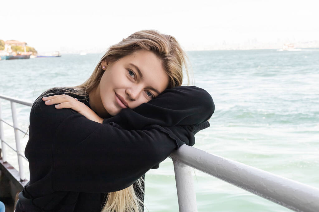 mujer joven rubia en suéter negro mirando a la cámara desde el ferry que cruza el estrecho del Bósforo - Foto, Imagen
