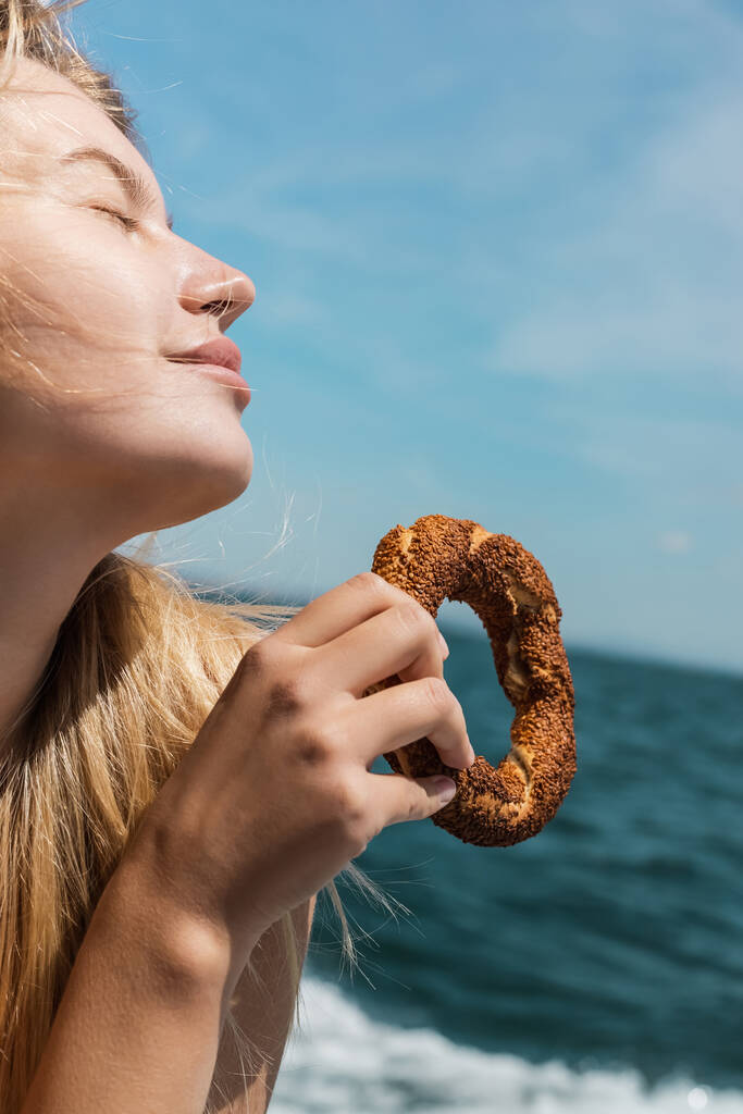 piacere donna bionda con gli occhi chiusi in possesso di bagel di sesamo turco vicino al mare  - Foto, immagini