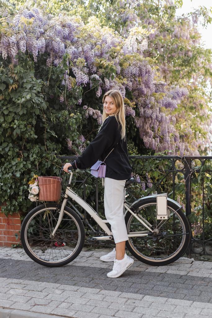 wesoła młoda kobieta w modnym stroju w pobliżu roweru i kwitnące drzewo w indyku - Zdjęcie, obraz