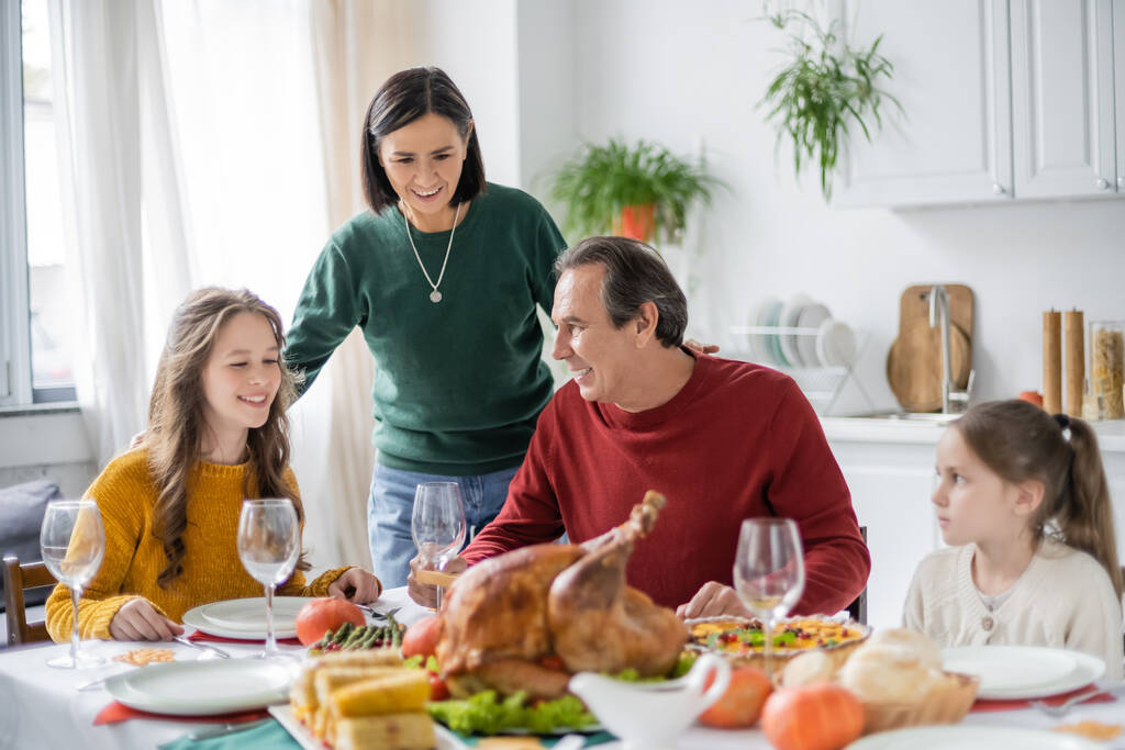 Smiling multiethnic grandparents and grandchildren sitting near blurred thanksgiving dinner at home  - 写真・画像