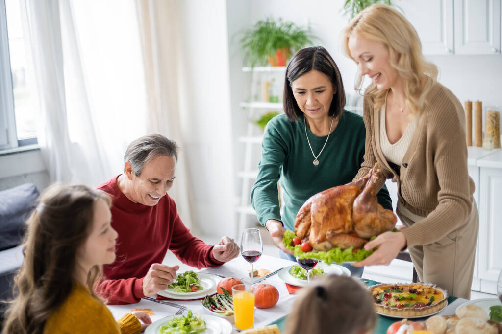 Többnemzetiségű nők kezében pulyka közel mosolygós család és hálaadási vacsora otthon  - Fotó, kép