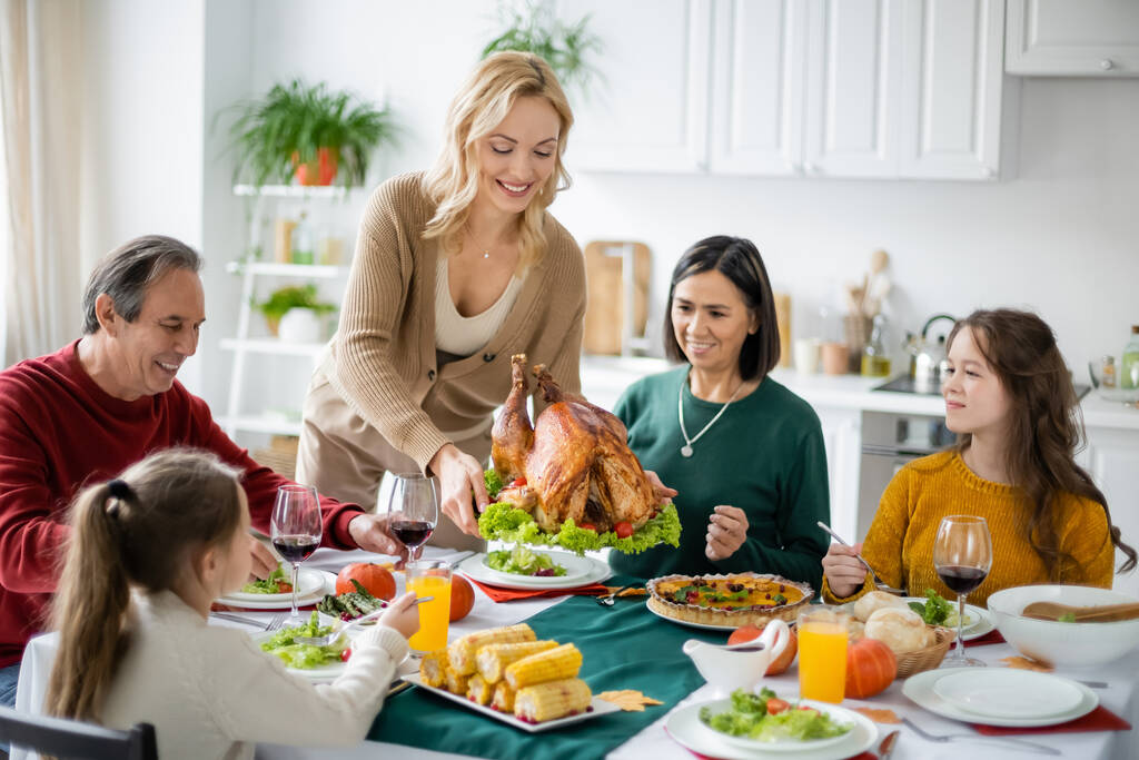 Mujer positiva sosteniendo pavo cerca de la familia multicultural y comida en casa  - Foto, imagen