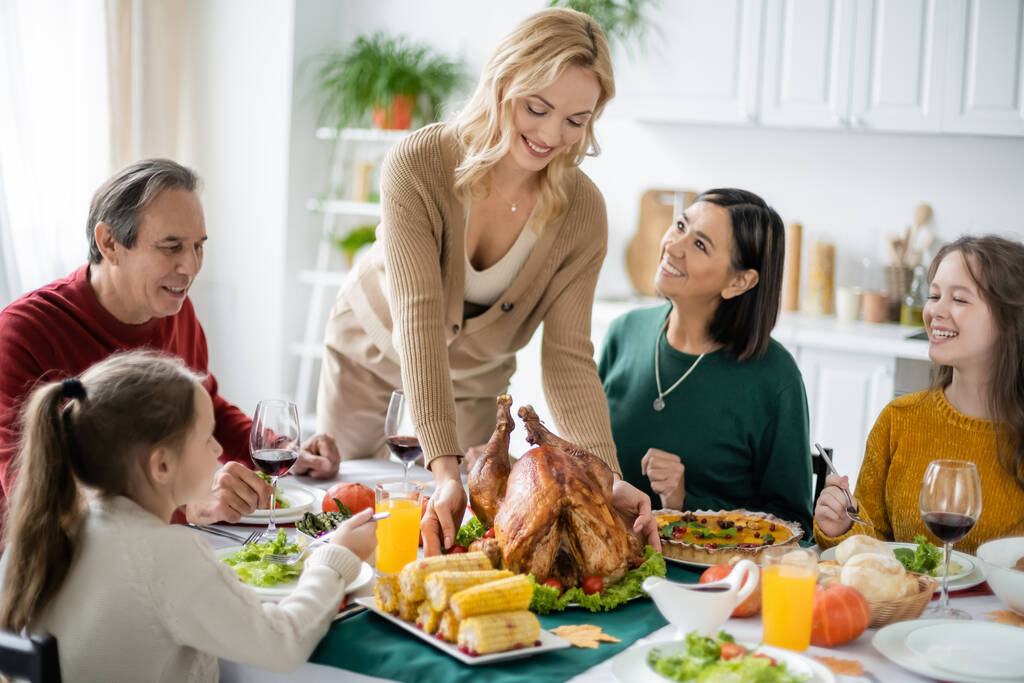 Sonriente familia multiétnica mirando a la mujer con sabroso pavo durante la cena de acción de gracias en casa  - Foto, imagen
