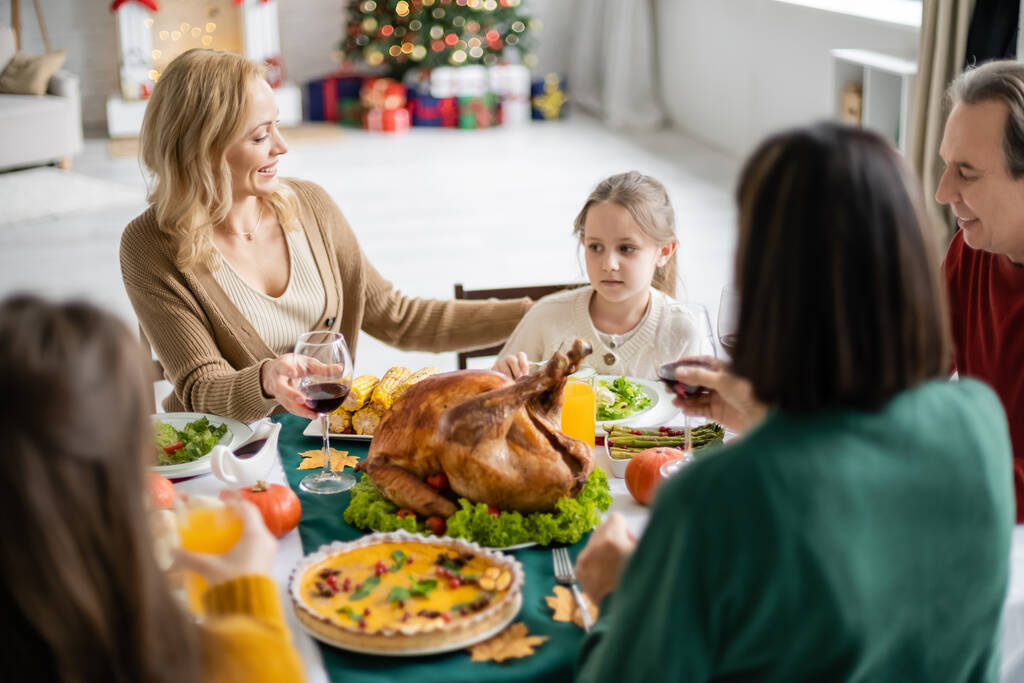 Улыбающаяся мать смотрит на ребенка рядом с размытой семьей и ужин в честь Дня Благодарения дома  - Фото, изображение