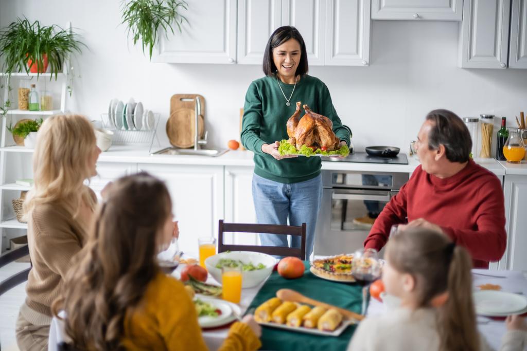 Positivo nonno multietnico con gustoso tacchino vicino alla famiglia offuscata durante la cena del Ringraziamento  - Foto, immagini