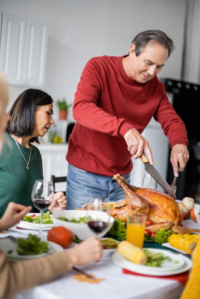 Hombre mayor cortando pavo cerca de la familia interracial durante la cena de Acción de Gracias en casa  - Foto, imagen