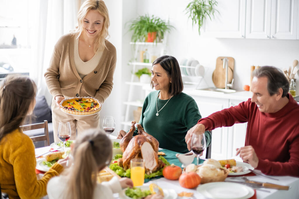 感謝祭の夕食の間に多文化の両親や子供の近くにパイを保持陽気な女性  - 写真・画像