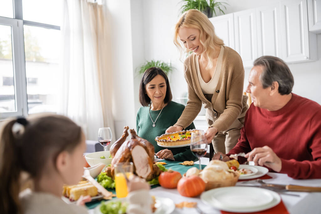 Pozitivní žena drží koláč v blízkosti multietnických rodičů a Díkůvzdání večeře doma  - Fotografie, Obrázek