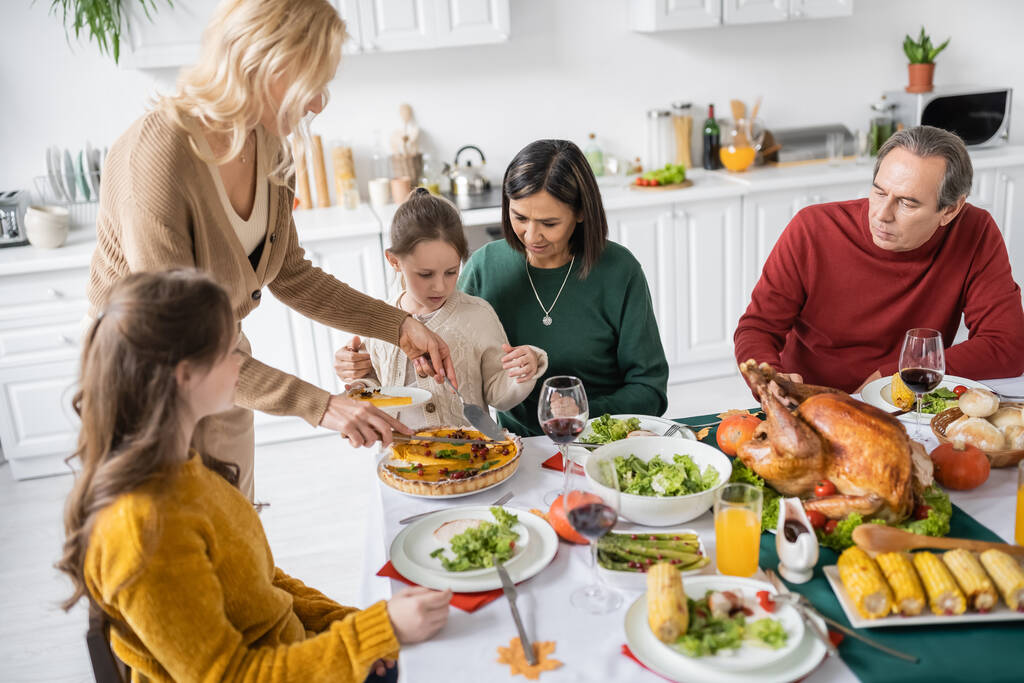 Мультикультурная семья с детьми, смотрящими на женщину, режущую пирог на День благодарения во время ужина дома  - Фото, изображение